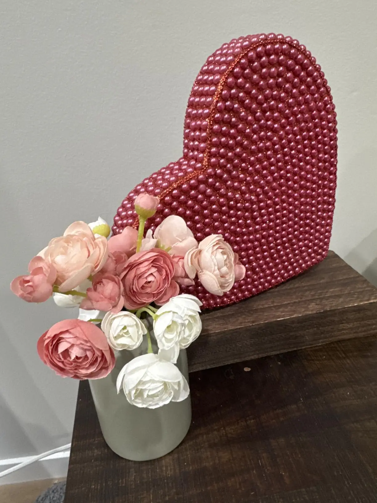 Dusty rose and white ranunculus arranged in a sage green vase beside a beaded heart decoration