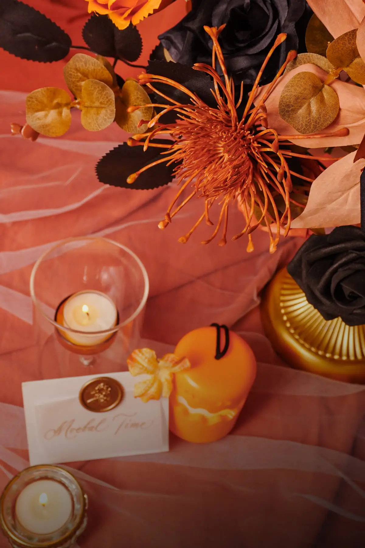 Top-down detail of a caramel orange artificial spider mum, brown eucalyptus and black rose leaves above a Halloween table with tea lights, a pumpkin candle and a Moefal Time place card